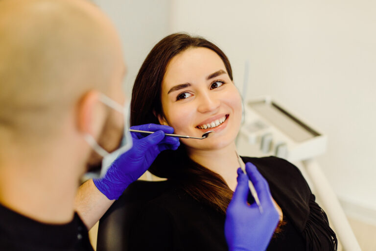 Dentist in blue gloves and mask performing an exam on a smiling patient prior to a crown lengthening consultation or cosmetic procedure.